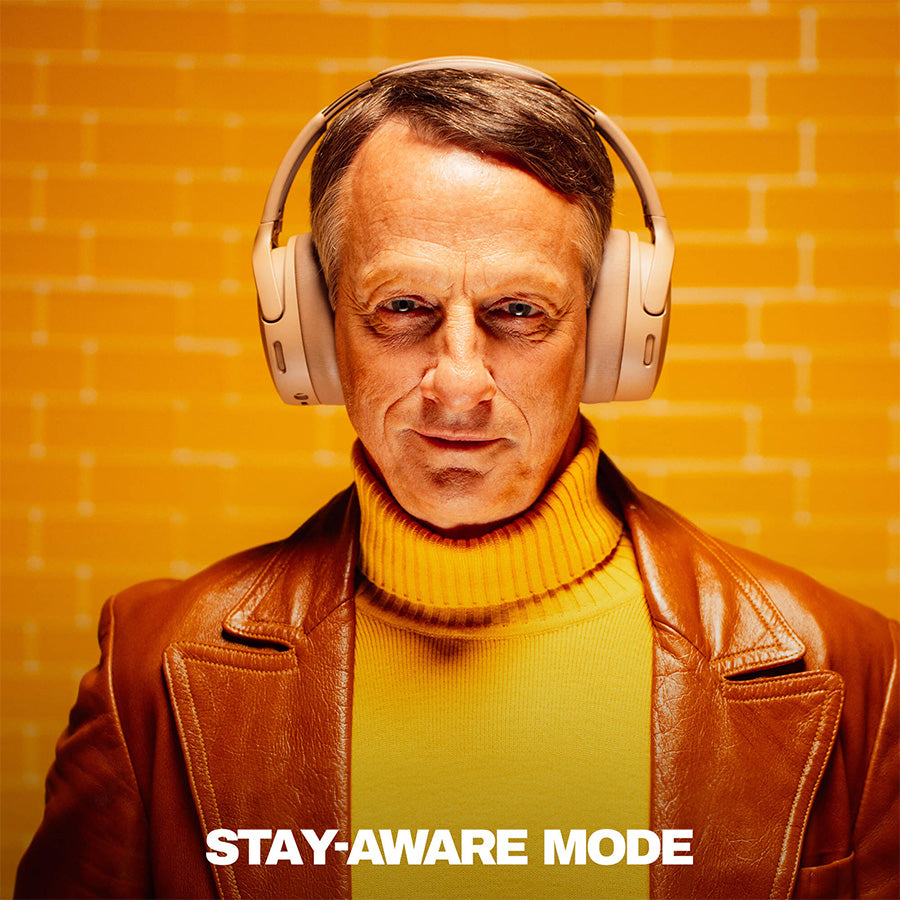 Tony Hawk wearing headphones with a text 'Adjustable Stay-Aware Mode' on an orange brick wall background | Primer