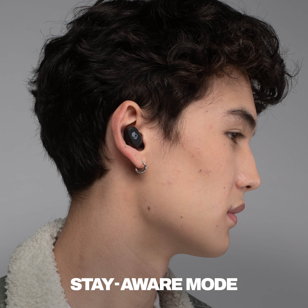 Person wearing a black wireless earbud with 'Stay-Aware Mode' text on a gray background | True Black