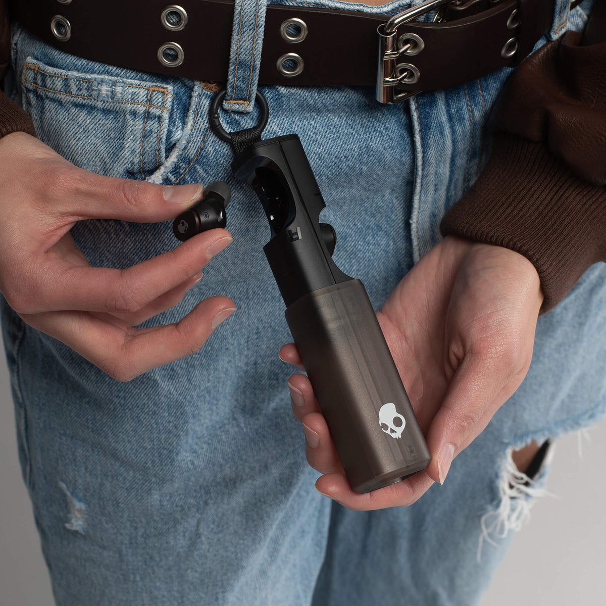 Person holding a black handheld device with a skull logo, wearing blue jeans and a brown jacket. | True Black