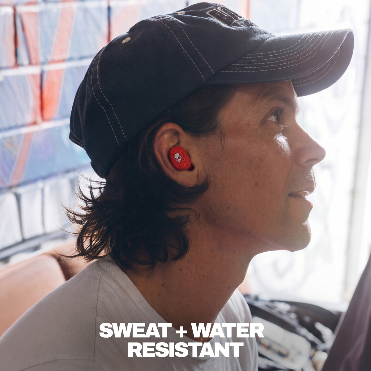 A man in profile wearing a black baseball cap and light gray t-shirt, with a red in-ear Dime 360 earbud visible in his ear. The text 'SWEAT + WATER RESISTANT' is overlaid at the bottom. | Skullcandy X Baker Skateboards