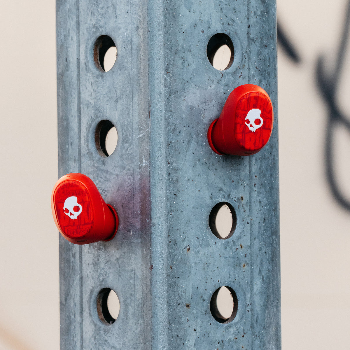 Two bright red Skullcandy X Baker Skateboards Dime 360 earbuds with white skull graphics mounted on a blue-gray metal rack. | Skullcandy X Baker Skateboards