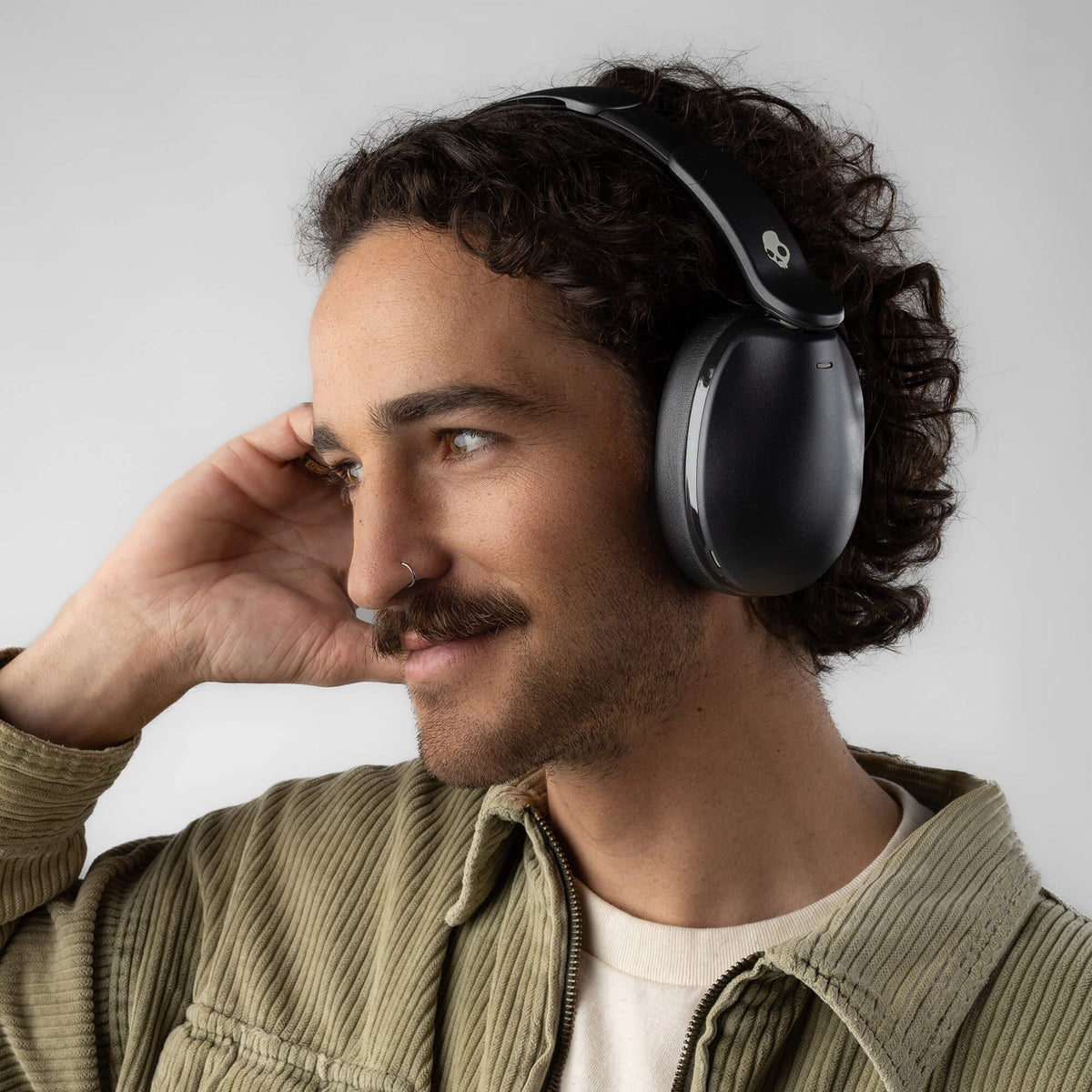 A man with curly dark hair and a mustache wearing black hesh 360 over-ear headphones and an olive green corduroy jacket, shown in a close-up profile against a light gray background. | True Black