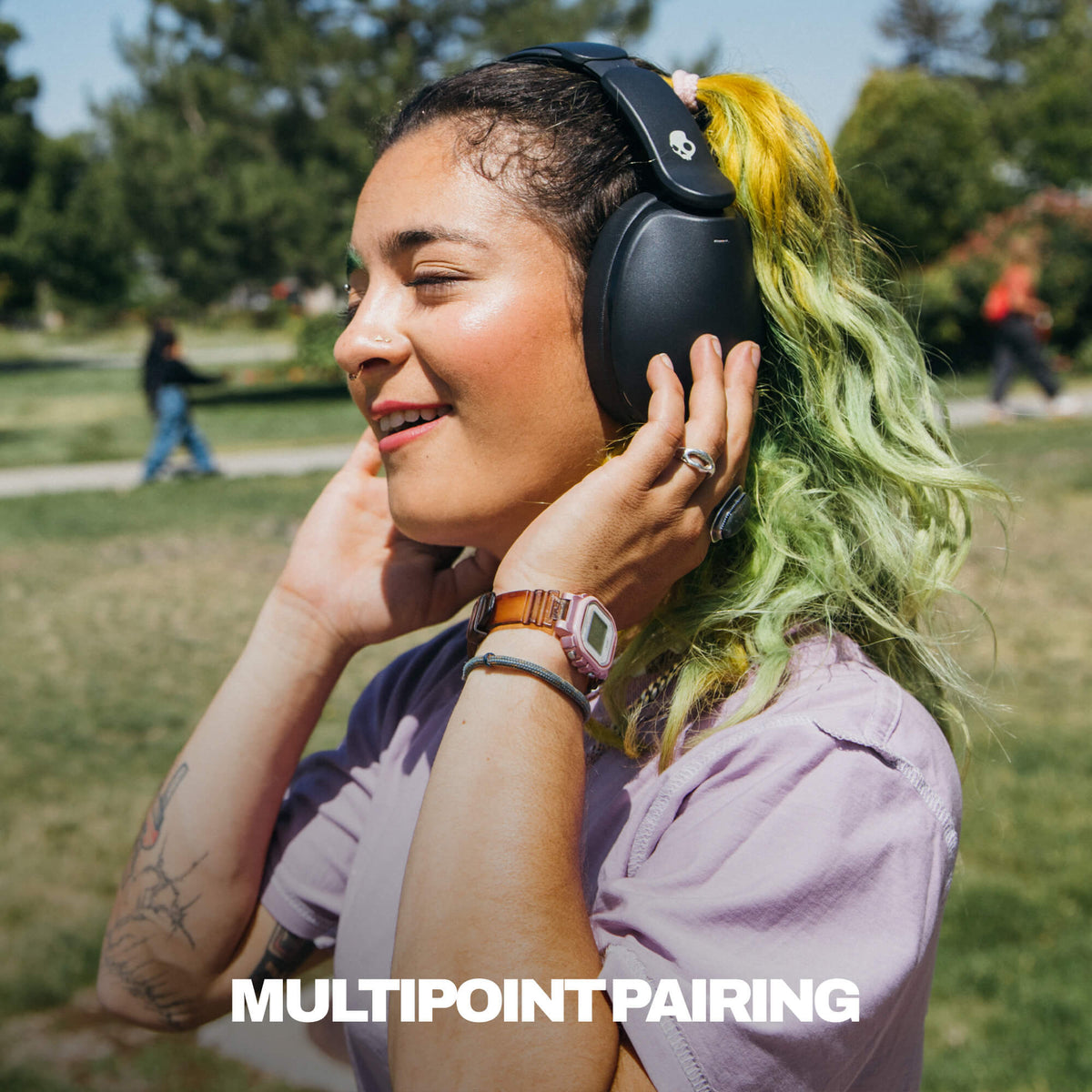 A woman outdoors in a park wearing black hesh 360 over-ear headphones and touching them with both hands. The text 'MULTIPOINT PAIRING' appears at the bottom of the image. | True Black