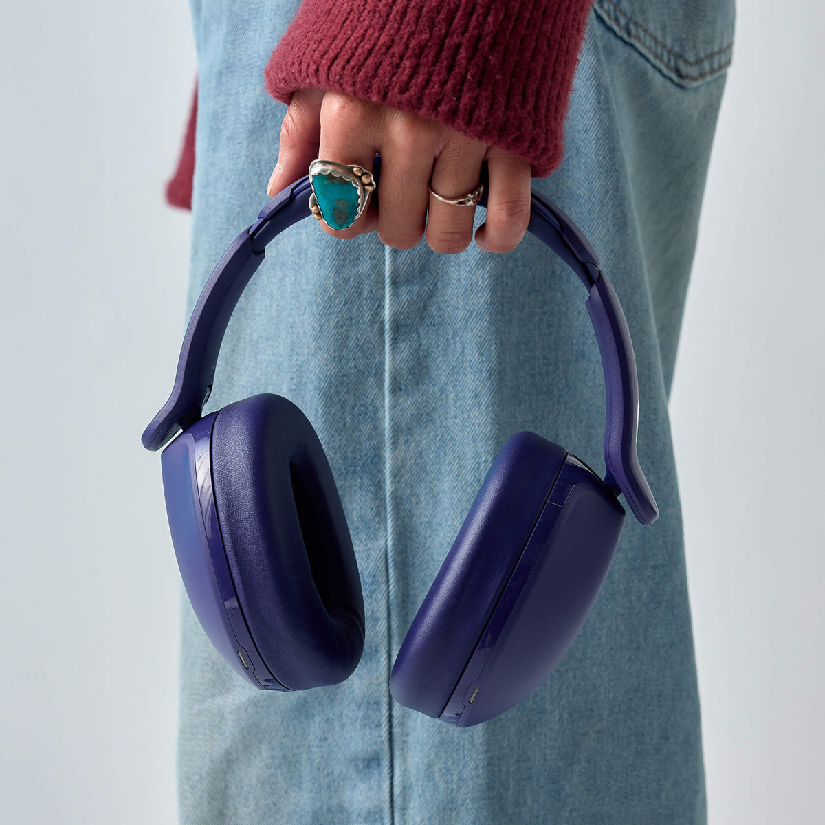 A person wearing a burgundy knit sweater and light blue denim jeans holds a pair of dark purple hesh 360 over-ear headphones. The person is wearing rings on their fingers, and the background is a clean, light gray studio backdrop. | Future