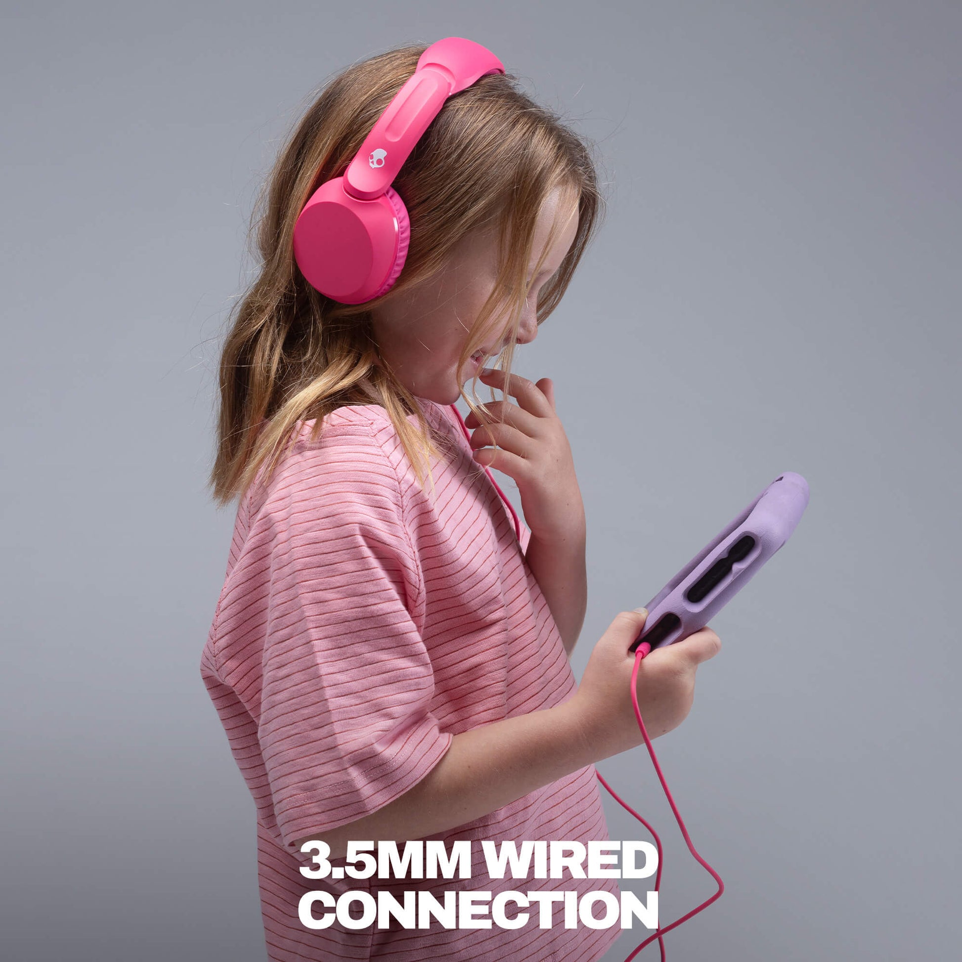A young girl with blonde hair wearing a pink striped shirt and bright pink over-ear headphones is shown from the side, holding a purple wired device with a red cable. The image has a gray studio background and white text at the bottom that reads '3. | Crazy Pink
