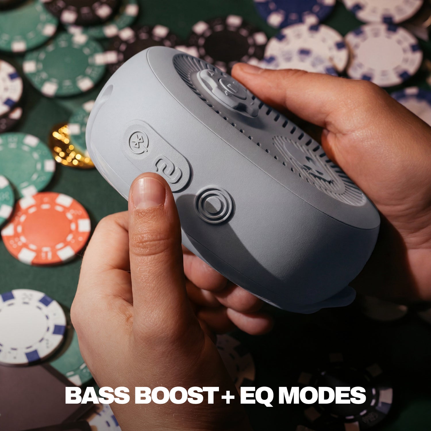 Person holding a gray speaker with buttons, surrounded by poker chips on a green felt surface. | Cement