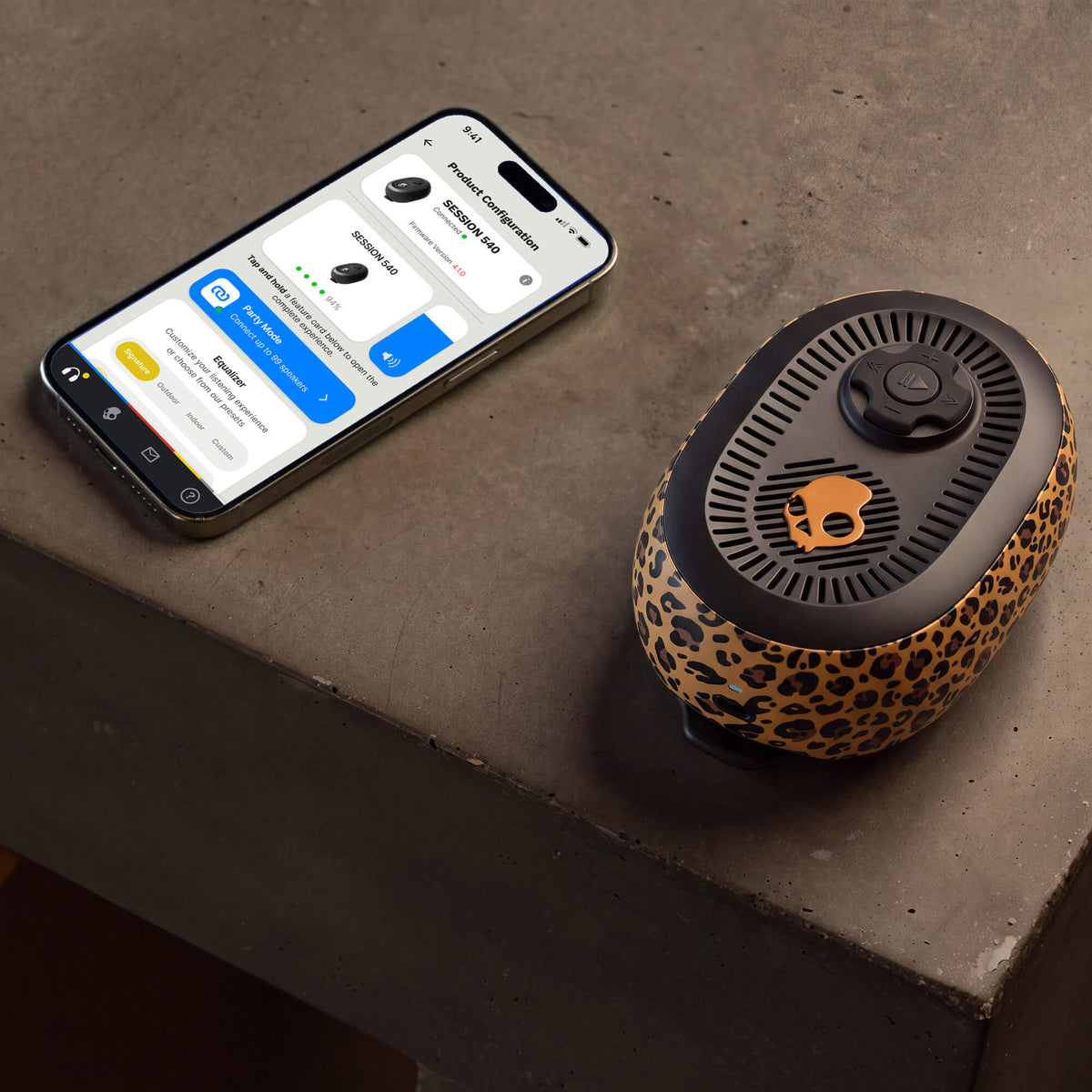 A smartphone displaying a companion app next to a session 540 speaker with a leopard-print pattern and black top grille, placed on a gray concrete surface. | Leopard