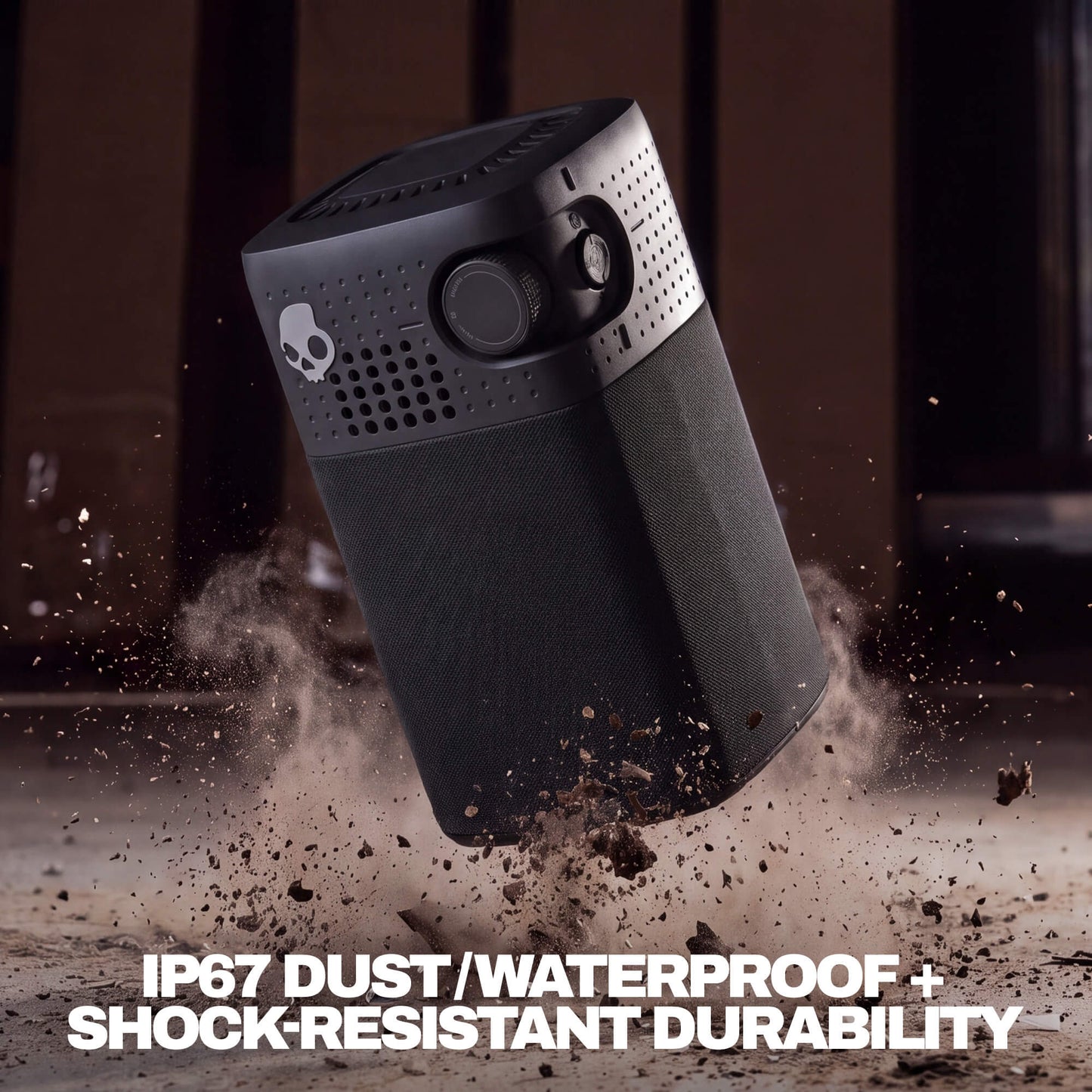 A rugged black session 900 speaker shown floating in midair with dirt and dust splashing around it. The product features a skull logo on the side and bold white text at the bottom reading 'IP67 DUST/WATERPROOF + SHOCK-RESISTANT DURABILITY'. | True Black