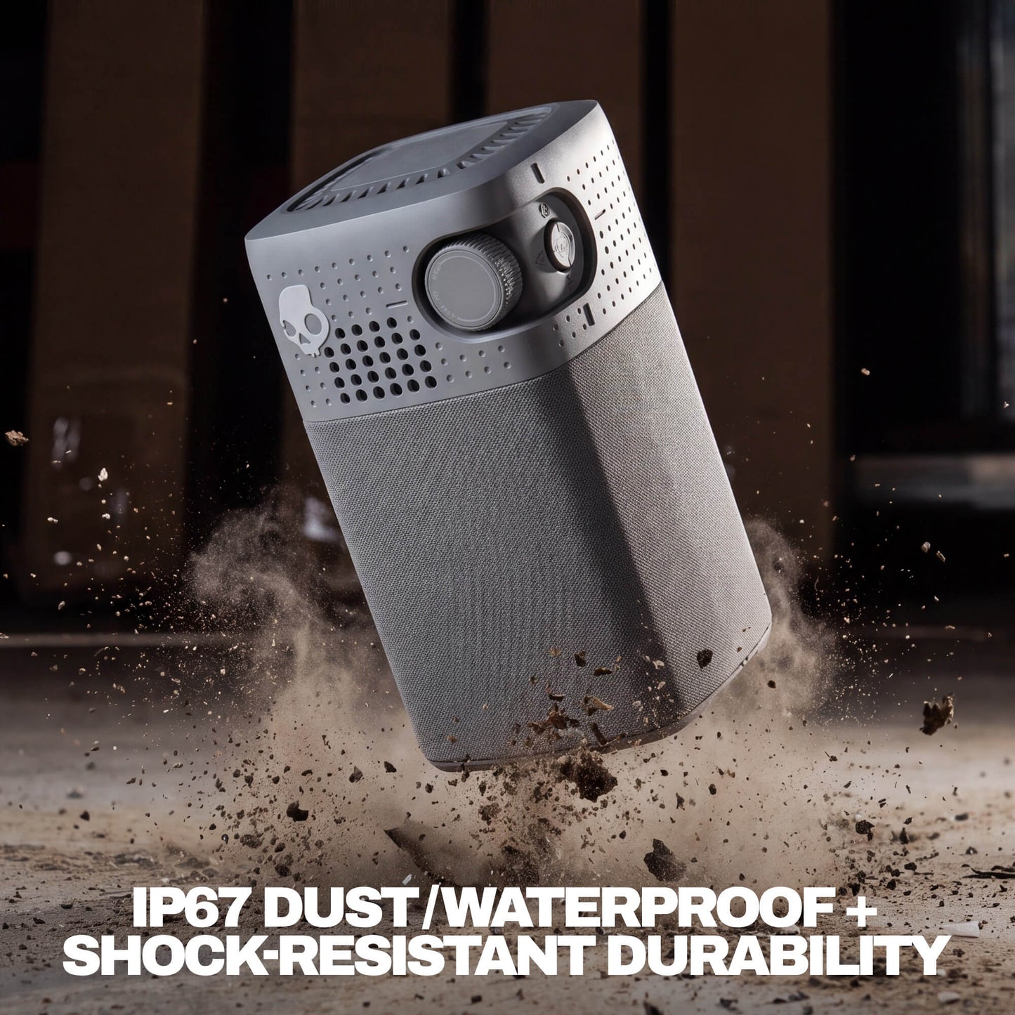 A gray session 900 portable speaker shown floating at an angle with dirt and dust particles around it, on a dark brown background. White text at the bottom reads 'IP67 DUST/WATERPROOF + SHOCK-RESISTANT DURABILITY'. | Cement