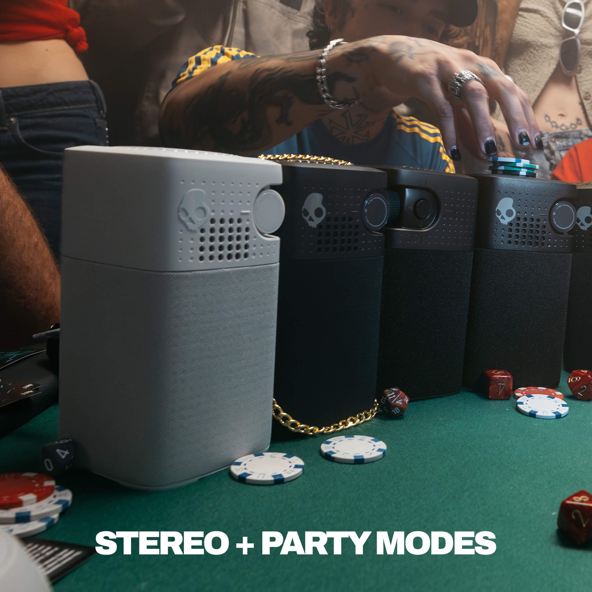 A group of session 900 speakers arranged on a green felt table with poker chips and a person's tattooed hand reaching toward them. The devices are shown in gray and black, with one gold chain draped across them. | Cement
