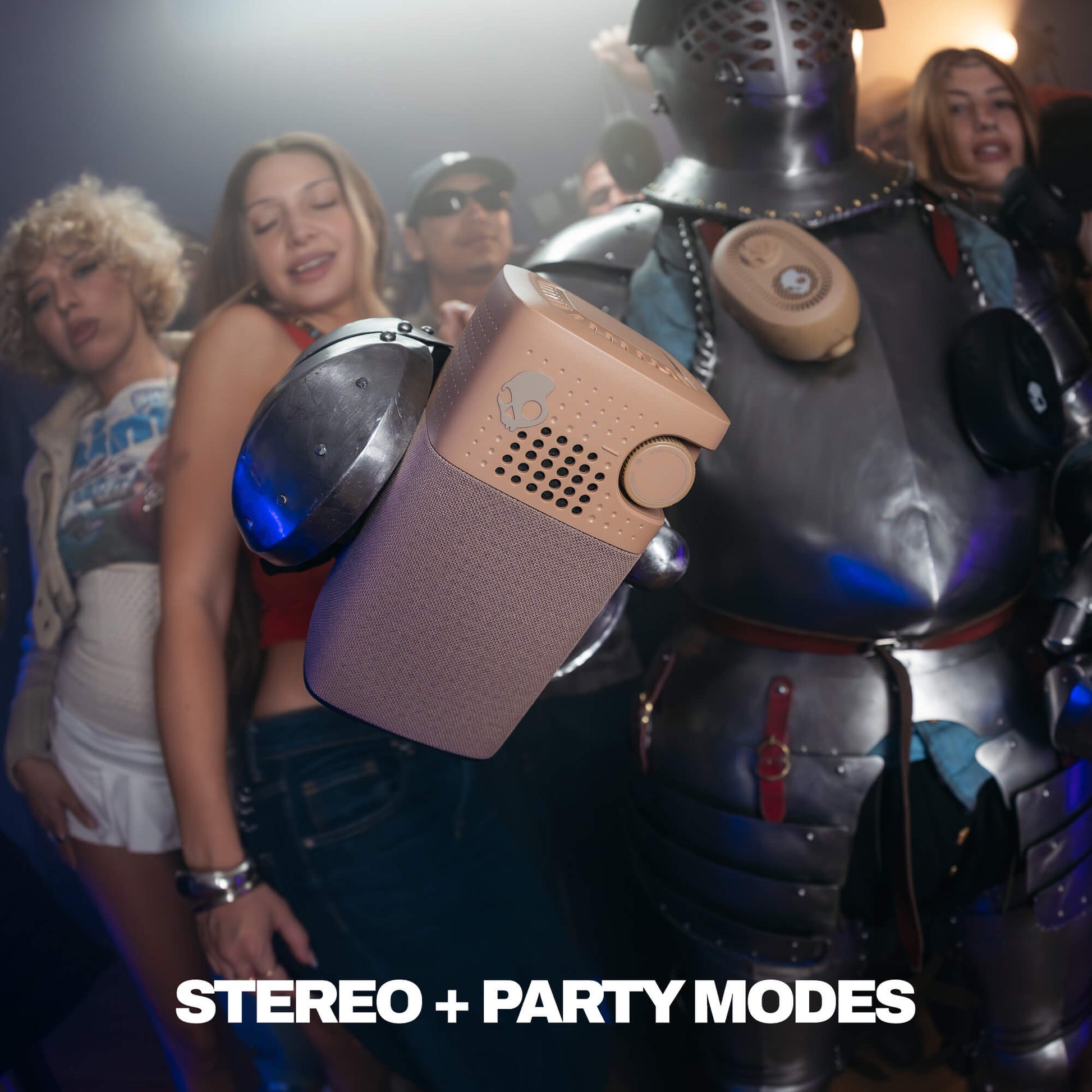 A portable Session 900 speaker with a textured taupe body is shown in a crowded party scene, with people and a knight in armor in the background. The text 'STEREO + PARTY MODES' is displayed at the bottom. | Primer
