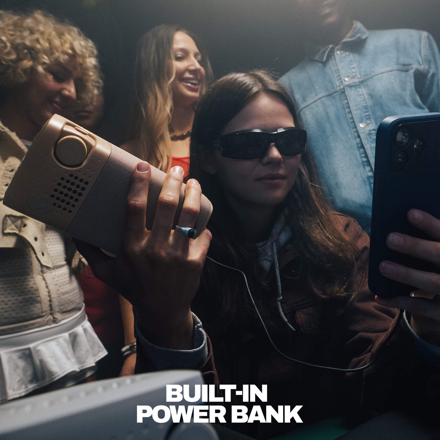A woman wearing dark sunglasses holds a session 900 speaker in one hand while taking a selfie with a smartphone. Other people are visible in the background, and the text 'BUILT-IN POWER BANK' appears at the bottom. | Primer