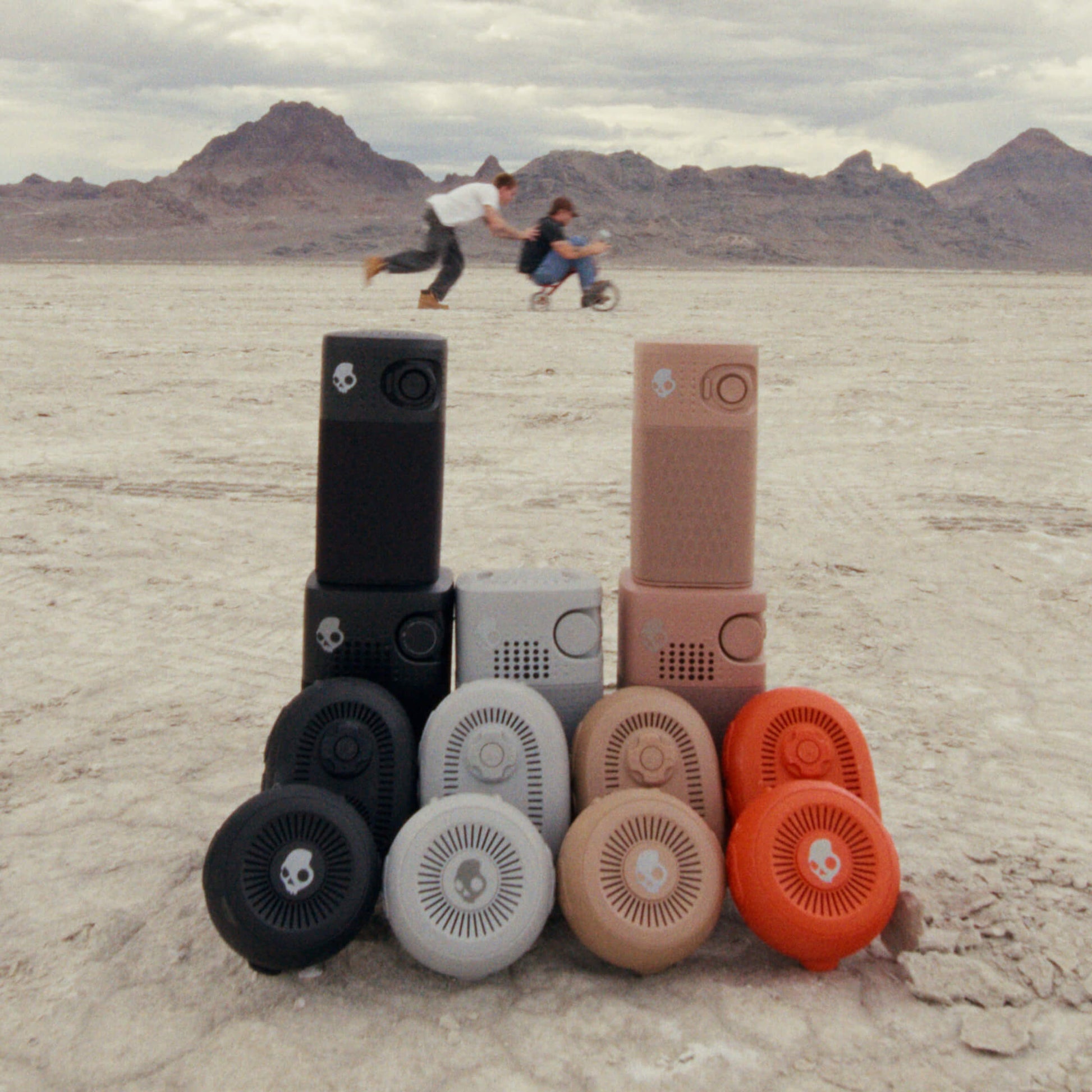 A set of portable speakers in multiple colors (black, gray, beige, orange) arranged in a pyramid on a desert flatland with mountains in the background. Two people are blurred in the background riding a bicycle. | Primer