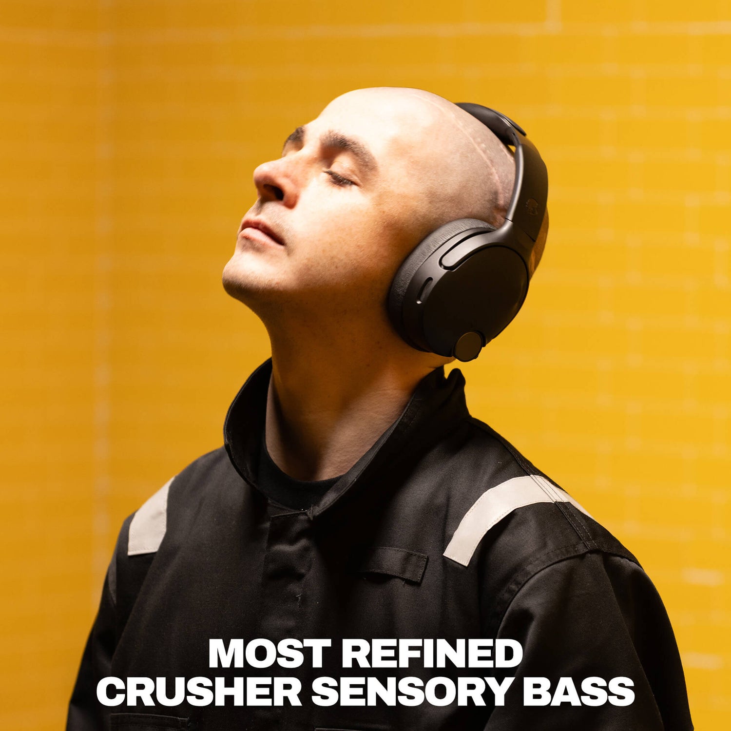 A man wearing black over-ear headphones and a black jacket with white shoulder stripes, looking upward against a bright yellow background. Large white text at the bottom reads 'MOST REFINED CRUSHER SENSORY BASS'.  | True Black