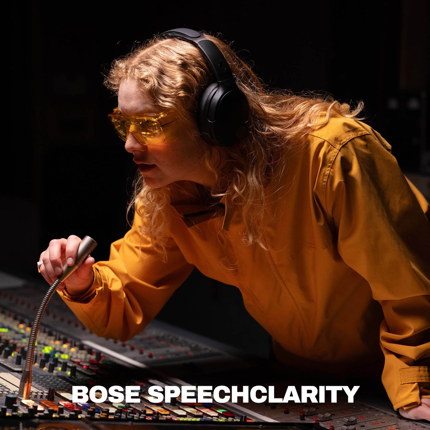 A woman in a recording studio wearing yellow-tinted safety glasses and black over-ear headphones, leaning over a mixing board and holding a microphone. The text 'BOSE SPEECHCLARITY' appears at the bottom. | True Black