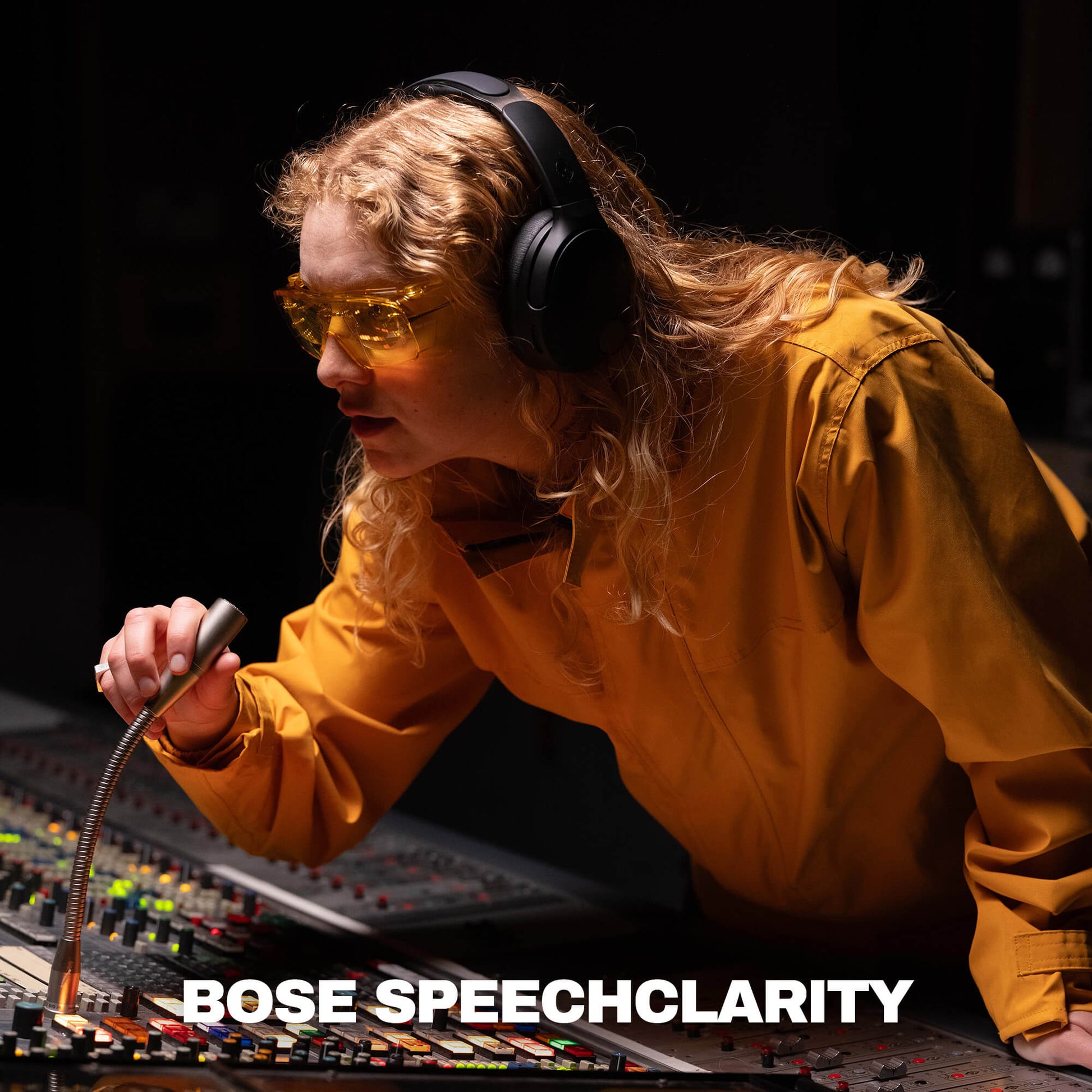 A woman in a recording studio wearing yellow-tinted safety glasses and black over-ear headphones, leaning over a mixing board and holding a microphone. The text 'BOSE SPEECHCLARITY' appears at the bottom. | True Black