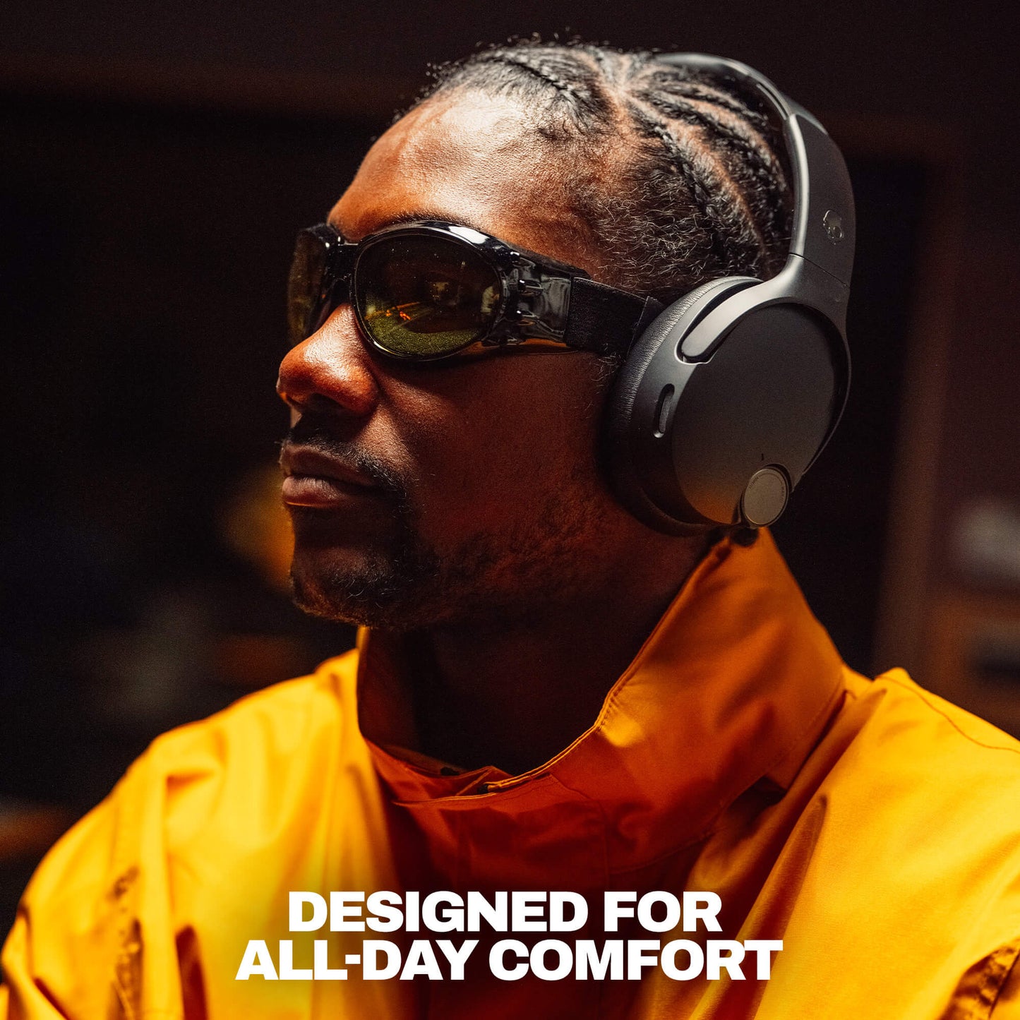 A man in a bright orange jacket wearing black over-ear headphones and dark sunglasses, shown in a dim indoor setting with the text 'DESIGNED FOR ALL-DAY COMFORT' at the bottom. | True Black