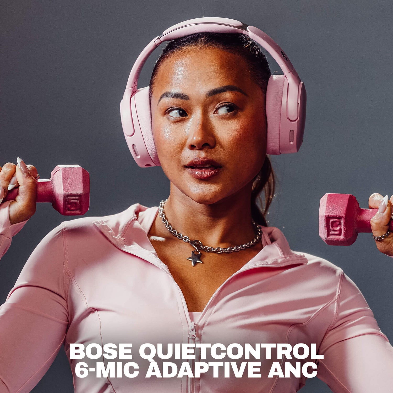 A woman wearing pink Bose QuietControl 6-MIC Adaptive ANC over-ear headphones and a matching pink athletic zip-up top, holding pink dumbbells on both sides against a dark gray studio background. White text at the bottom reads 'BOSE QUIETCONTROL 6-MIC ADAPTIVE ANC'. | Candy