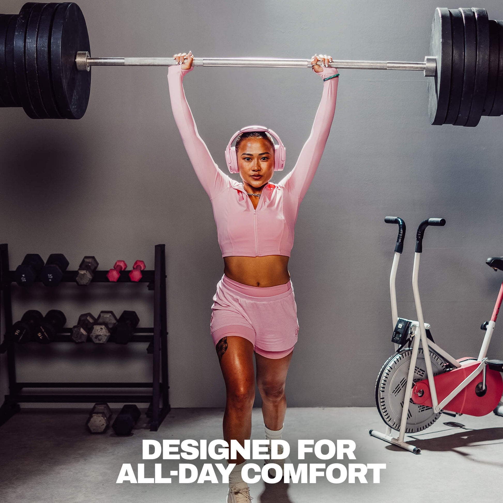 A woman in a pink athletic zip-up hoodie and matching shorts lifts a barbell overhead in a gym setting. She is wearing pink headphones, with dumbbells on a rack to her left and a stationary bike to her right. | Candy