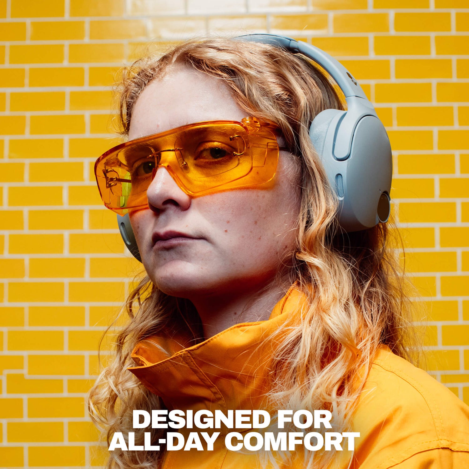 A woman wearing light blue over-ear headphones and translucent orange-tinted safety glasses, set against a yellow brick wall background with the text 'DESIGNED FOR ALL-DAY COMFORT' at the bottom. | Cement