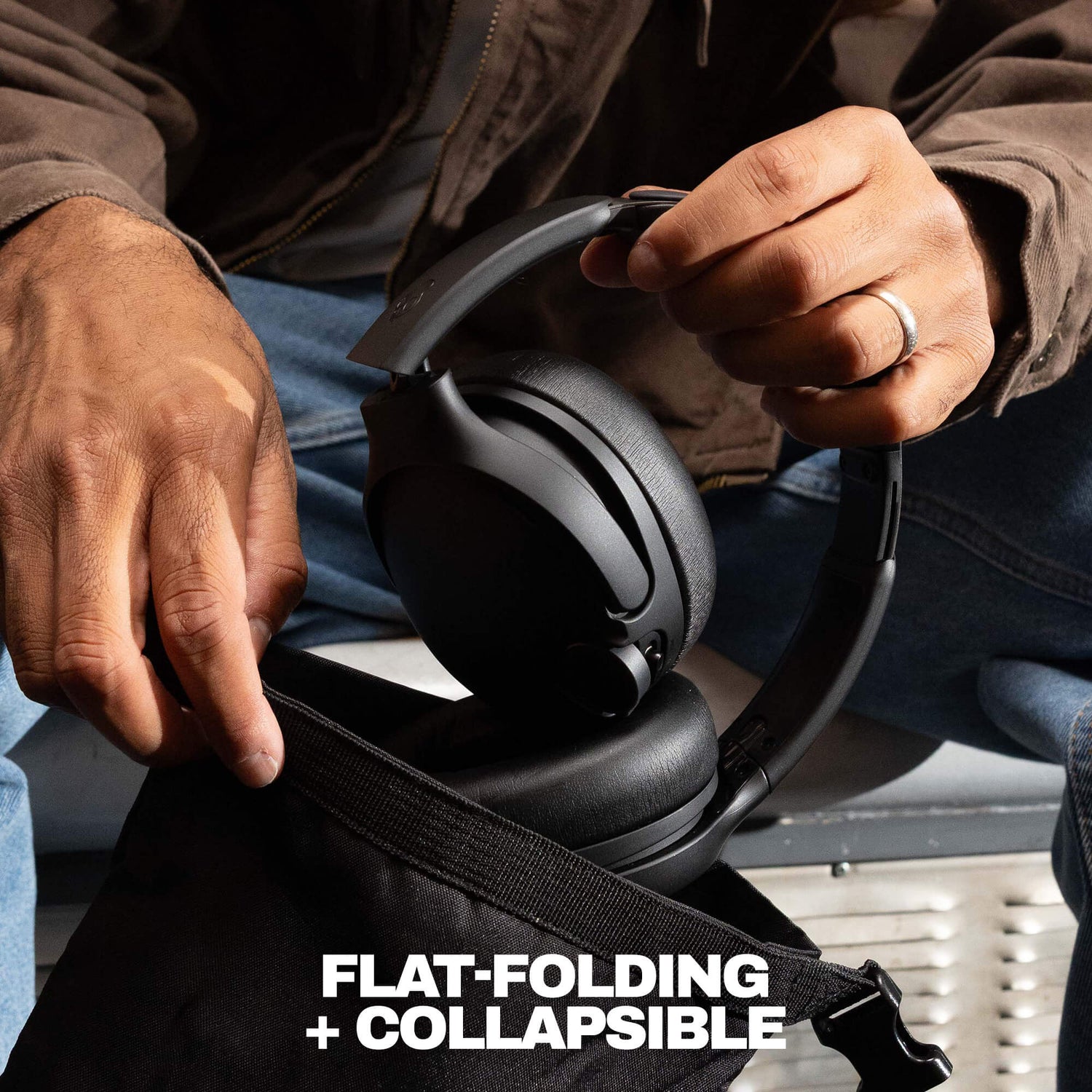 A close-up of a person holding a black over-ear headset over an open black bag, with the text 'FLAT-FOLDING + COLLAPSIBLE' at the bottom. The person is wearing a brown jacket and blue jeans.| Cement