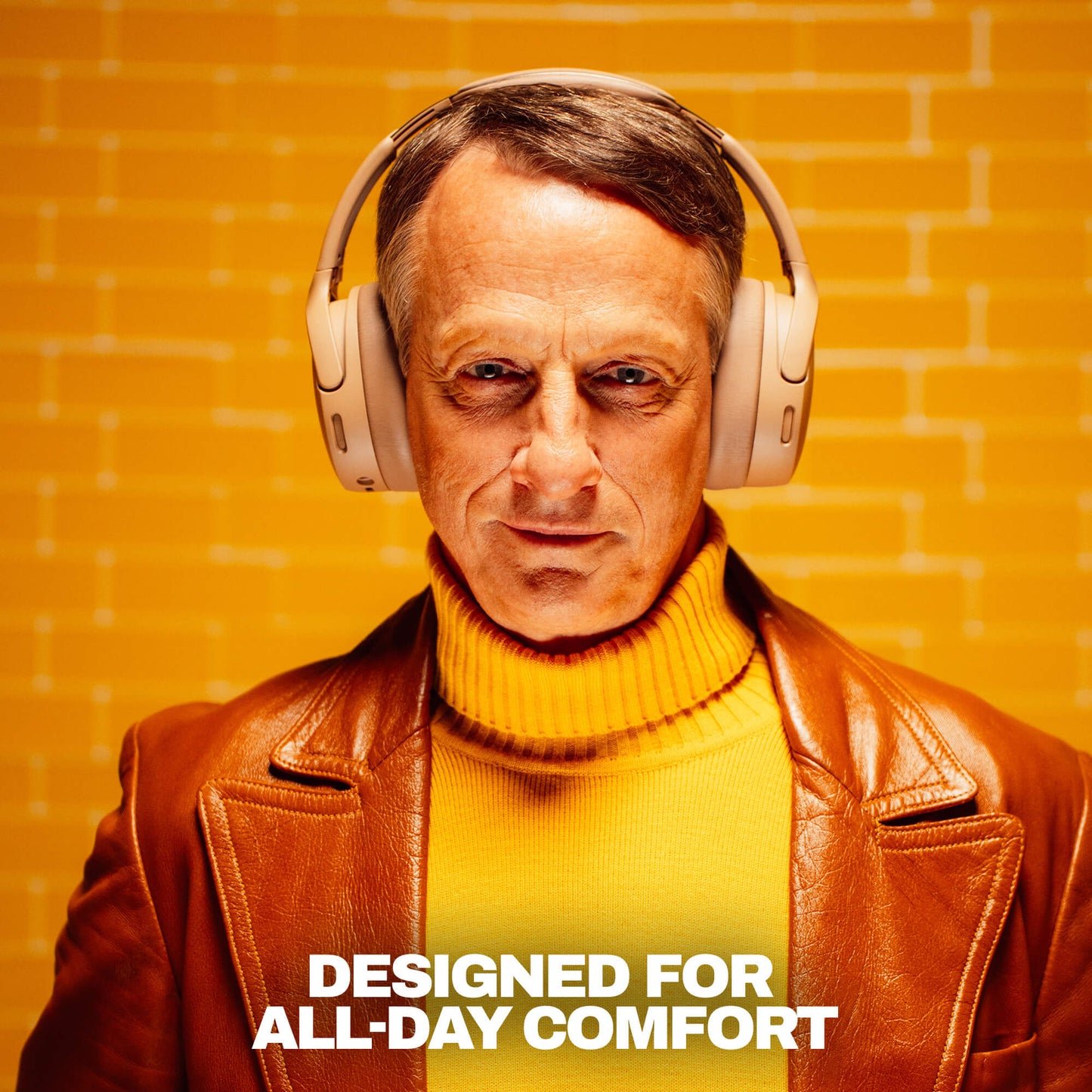 A man wearing beige over-ear headphones, a brown leather jacket, and a yellow turtleneck sweater stands against a yellow brick wall background with the text 'DESIGNED FOR ALL-DAY COMFORT' at the bottom. | Primer