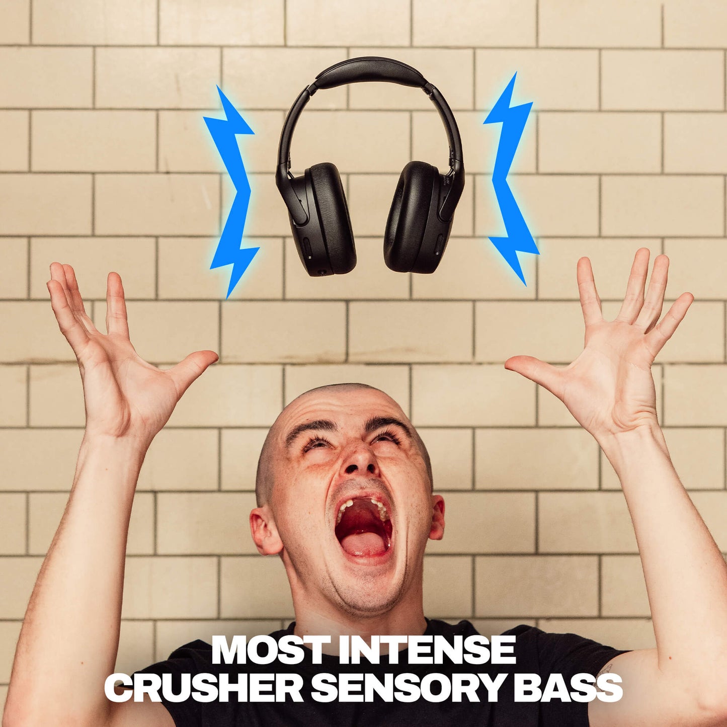 A man with his hands raised in front of a beige tiled wall, looking upward with an open mouth, with a pair of black Crusher 720 over-ear headphones floating above him and blue lightning bolts indicating sound. White text at the bottom reads 'MOST INTENSE CRUSHER SENSORY BASS'. | True Black