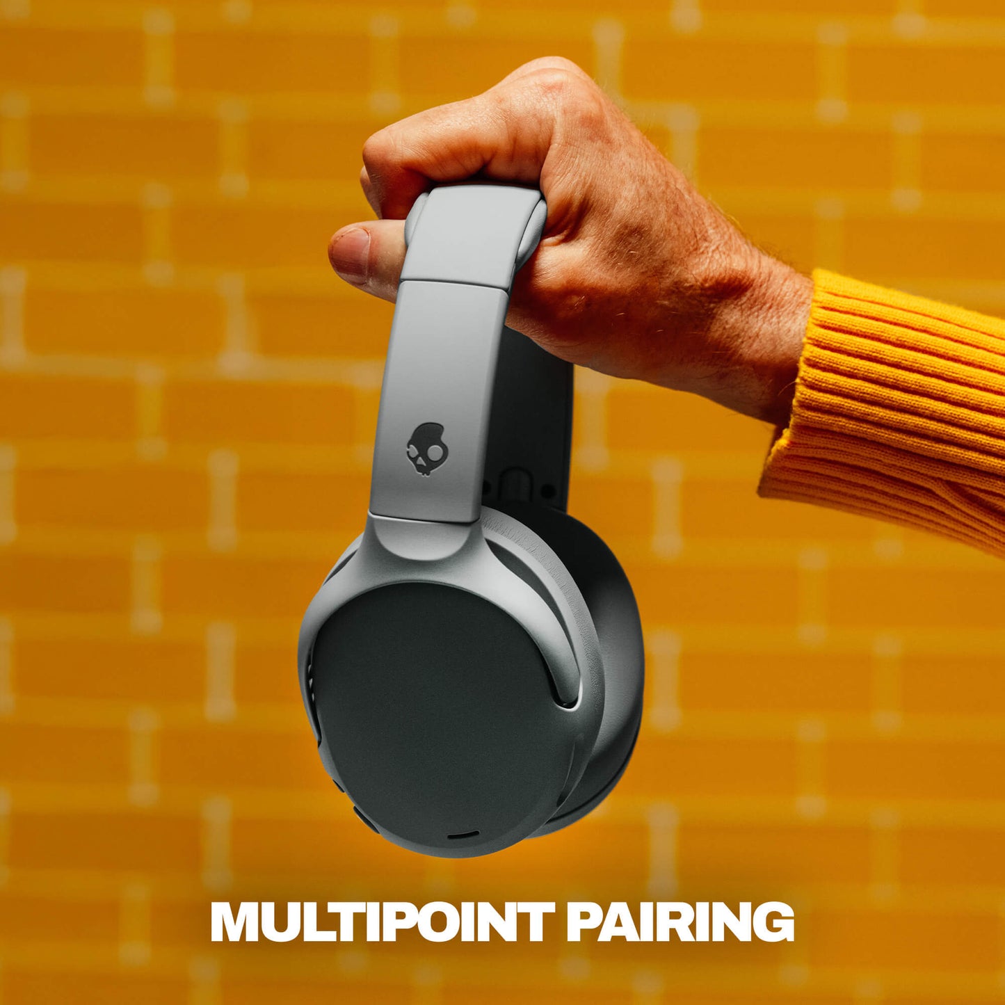 Hand holding a pair of headphones with 'MultiPoint Pairing' text on a yellow brick wall background | Cement