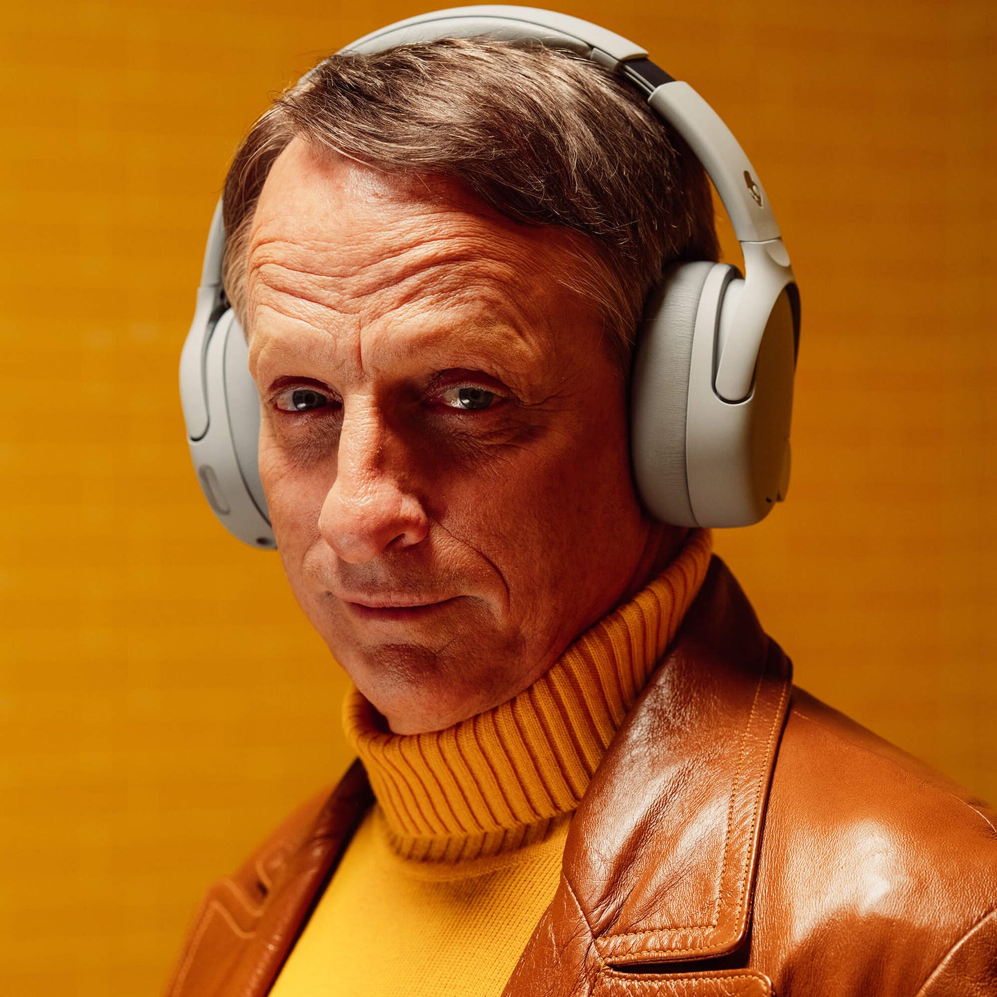 Tony Hawk wearing Crusher 720 headphones and a brown leather jacket against an orange background | Cement