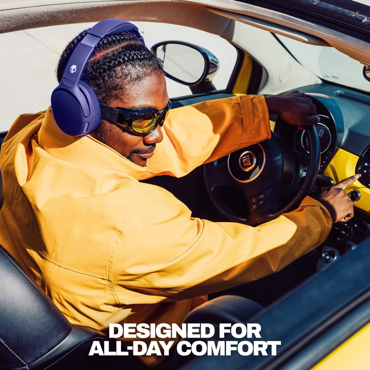 A man wearing Crusher 720 deep blue over-ear headphones and sunglasses sits in the driver's seat of a convertible car, in a mustard yellow jacket. The text 'DESIGNED FOR ALL-DAY COMFORT' is displayed at the bottom. | Future