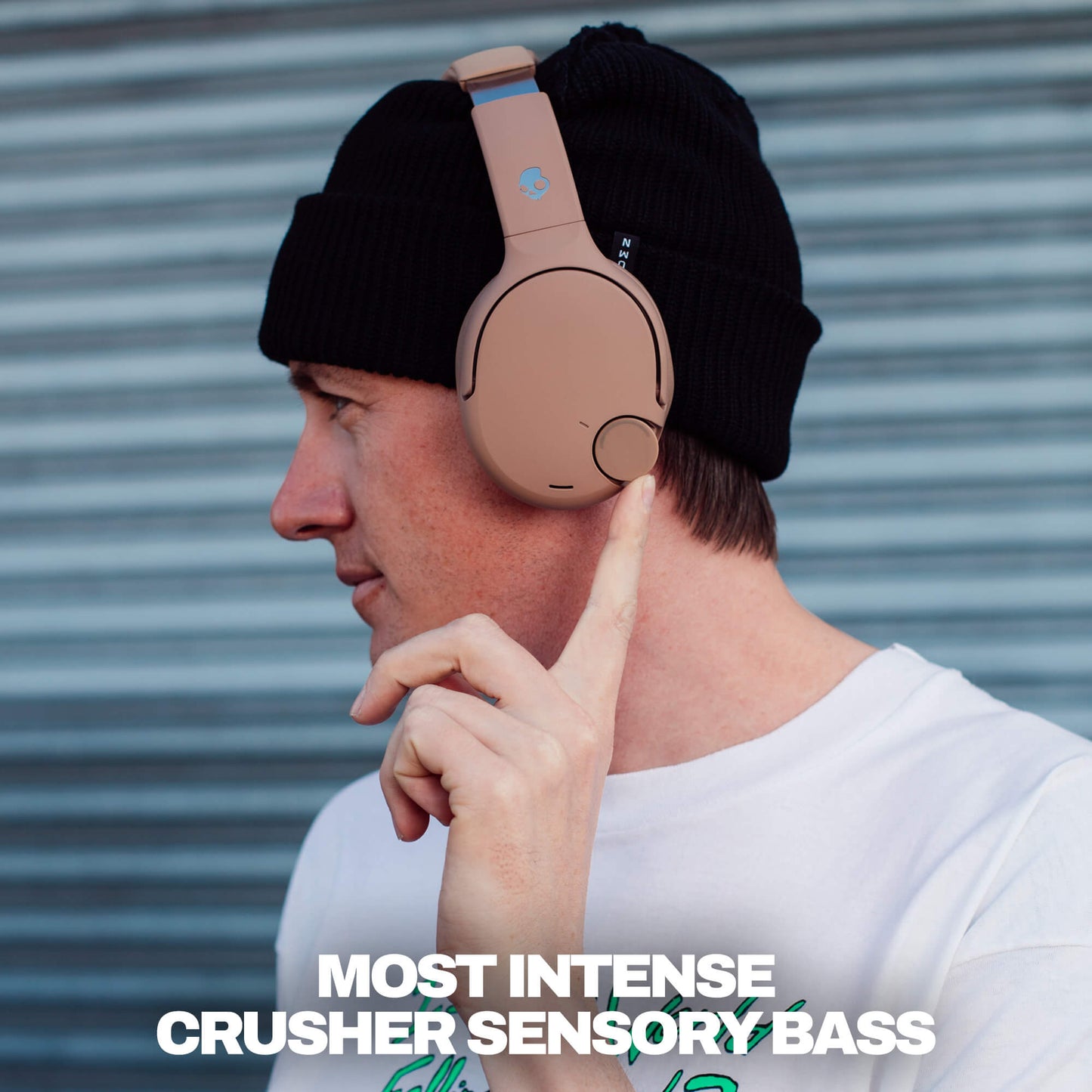Man wearing Crusher 720 headphones over a black beanie looking sideways and 'MOST INTENSE CRUSHER SENSORY BASS' text against a stripe textured blue metal wall. | Primer