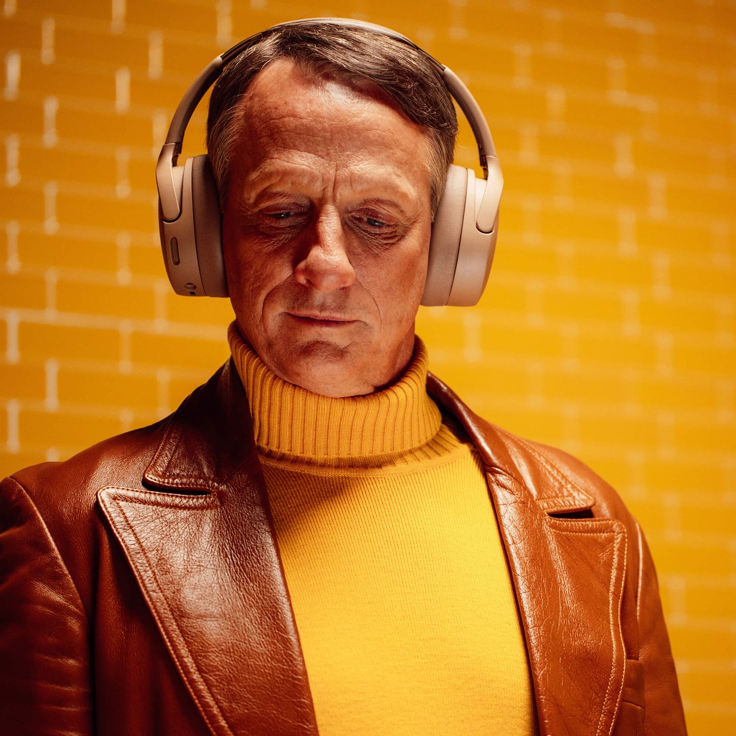 Tony Hawk wearing Crusher 720 headphones and a brown leather jacket over a yellow turtleneck against a yellow brick wall. | Primer